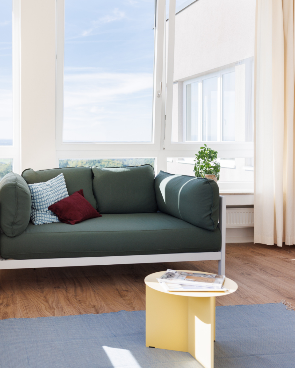 Modern living area in a POHA House apartment featuring a dark green sofa, yellow side table, and large window with an open pane overlooking the sky.