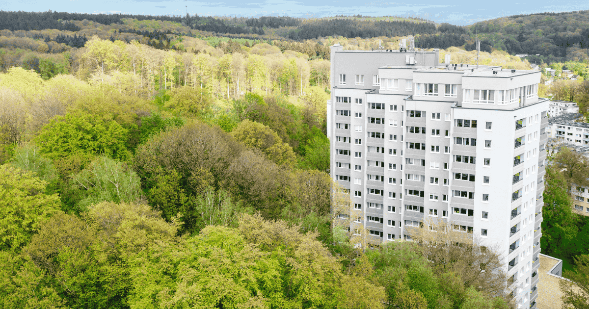 A tall, modern apartment building, POHA House Preuswald, stands amidst a lush green forest.