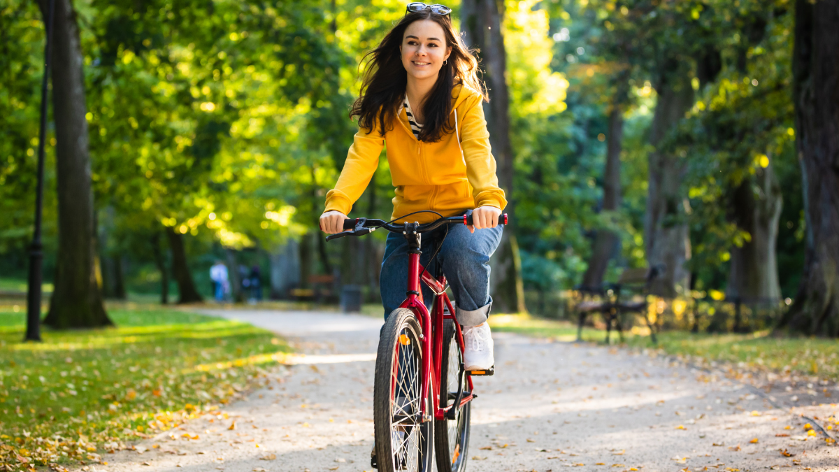 Young woman with long dark hair, wearing a yellow hoodie and blue jeans, happily riding a bicycle down a tree-lined, sun-dappled path in a park.
