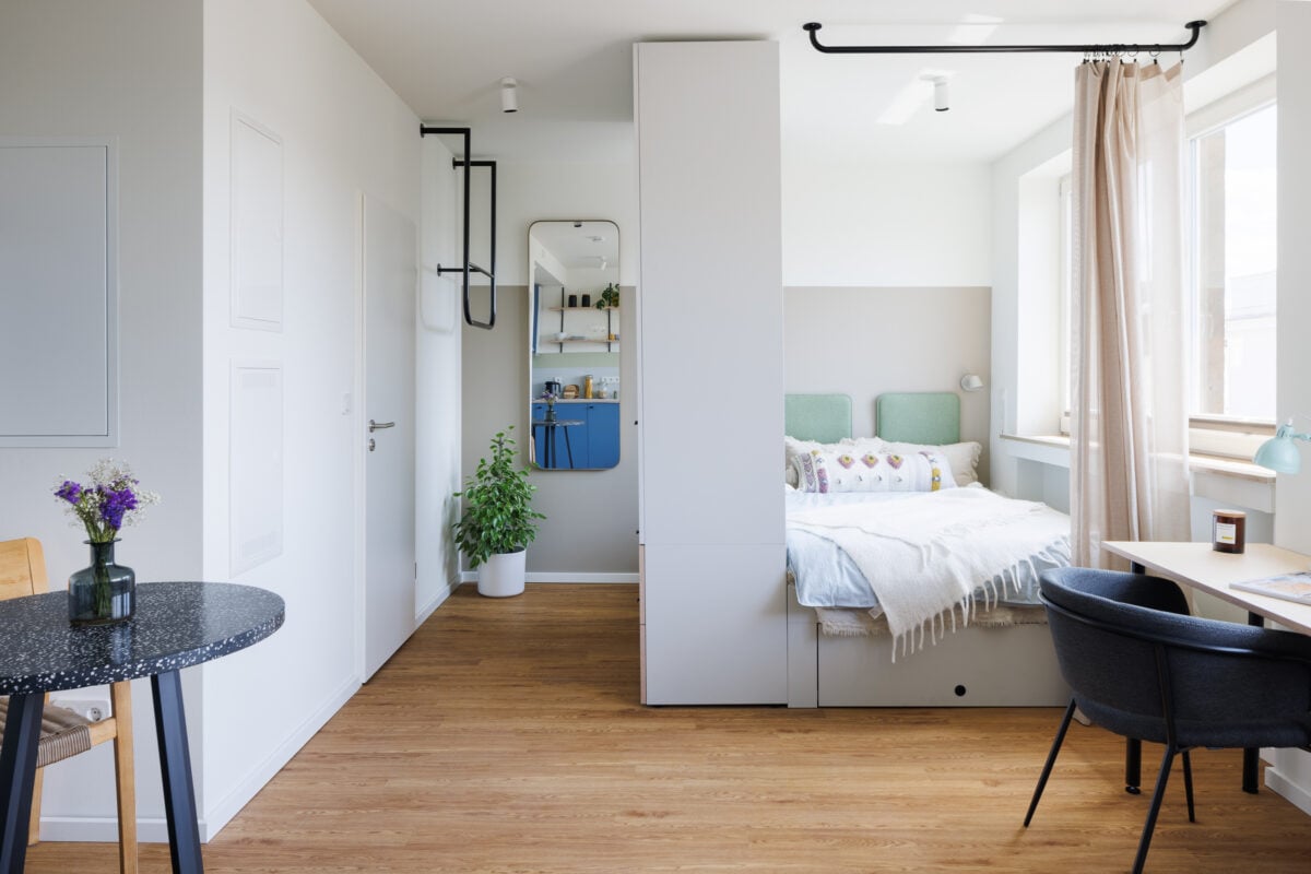 Interior view of a modern, minimalist studio apartment at POHA House Aachen featuring a smart, space-saving layout. The room includes light wood flooring, white walls, and a large central wardrobe that acts as a room divider, partially concealing a cozy bed nook with mint green headboards and a tasseled throw. To the left, a small dining area features a dark speckled bistro table with purple flowers; to the right, a dedicated workspace sits by a bright window. A tall mirror reflects a vibrant blue kitchenette in the background, enhancing the sense of open space.