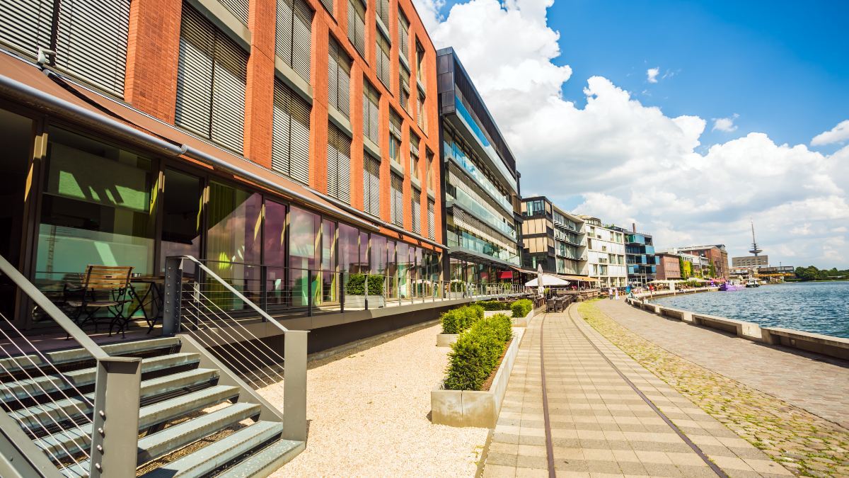 Modern architecture and red brick buildings lining the waterfront promenade of the Münster harbor (Hafen) in Germany on a bright, sunny day.