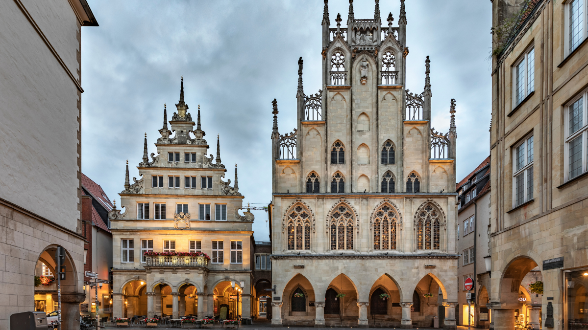 The historic Gothic architecture of the Münster City Hall (Rathaus) and the adjacent late Renaissance buildings, illuminated at dusk in the city center of Münster, Germany.