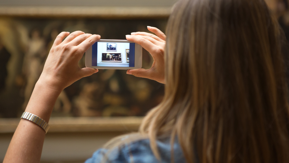 A person with long blonde hair taking a picture of an artwork using a white smartphone in a museum or art gallery. A classic painting is blurred in the background.