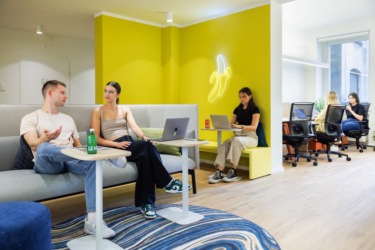 Young adults working and talking in a colorful modern office. A man and woman chat on a gray sofa while another woman works at a small table against a bright yellow wall with a neon banana sign.