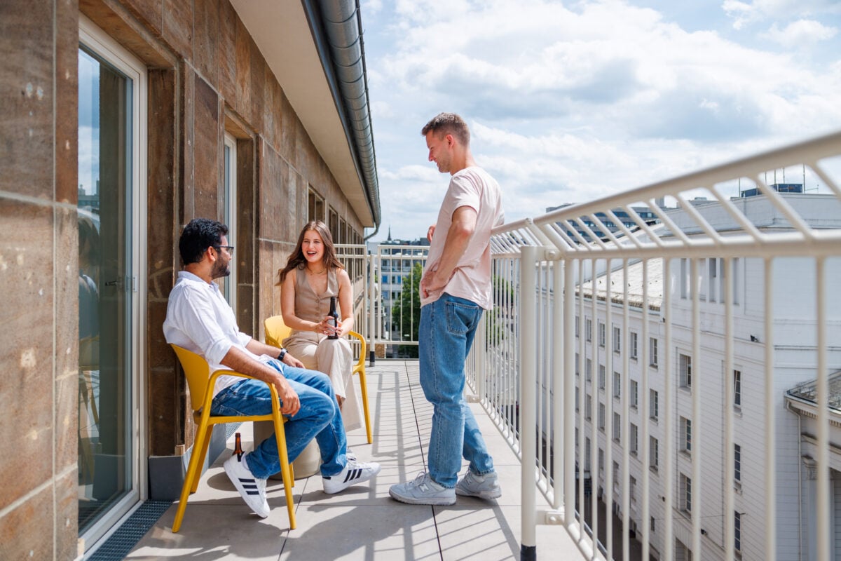 Three young adults are socialising on a sunny, spacious rooftop terrace at POHA House Aachen. Two individuals sit in bright yellow chairs against a textured stone wall, while a third person leans casually against the white metal railing. The terrace offers a view of the surrounding white urban architecture under a bright, slightly cloudy sky, capturing a relaxed community atmosphere