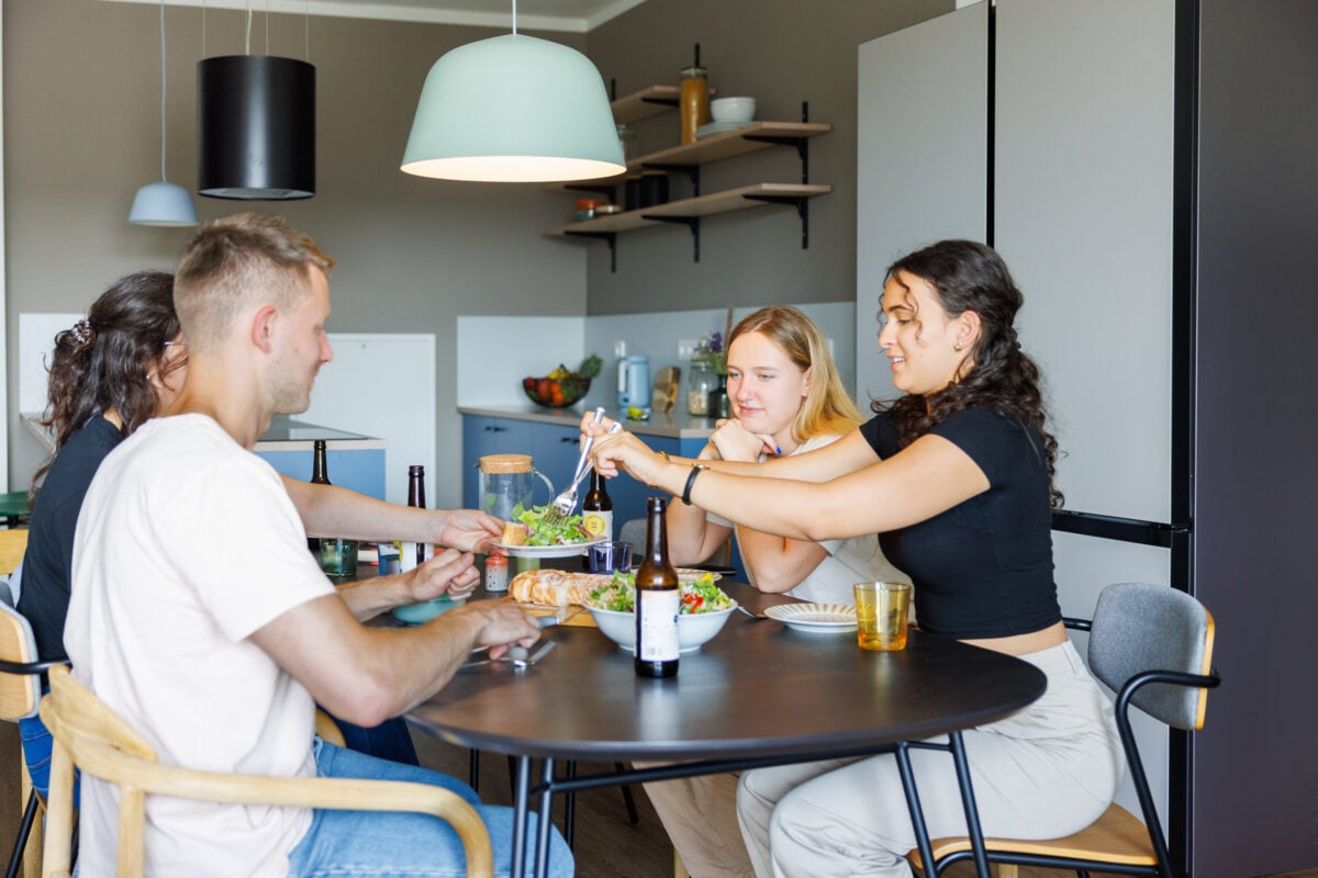 A group of four young adults is sharing a meal around a dark oval dining table in a modern, open-plan kitchen at POHA House Aachen. One person serves salad onto a plate while others look on and engage in conversation. The background features a stylish kitchen with blue cabinetry, open wooden shelving, and a large mint-green pendant light hanging above the table, creating a warm and communal living atmosphere.
