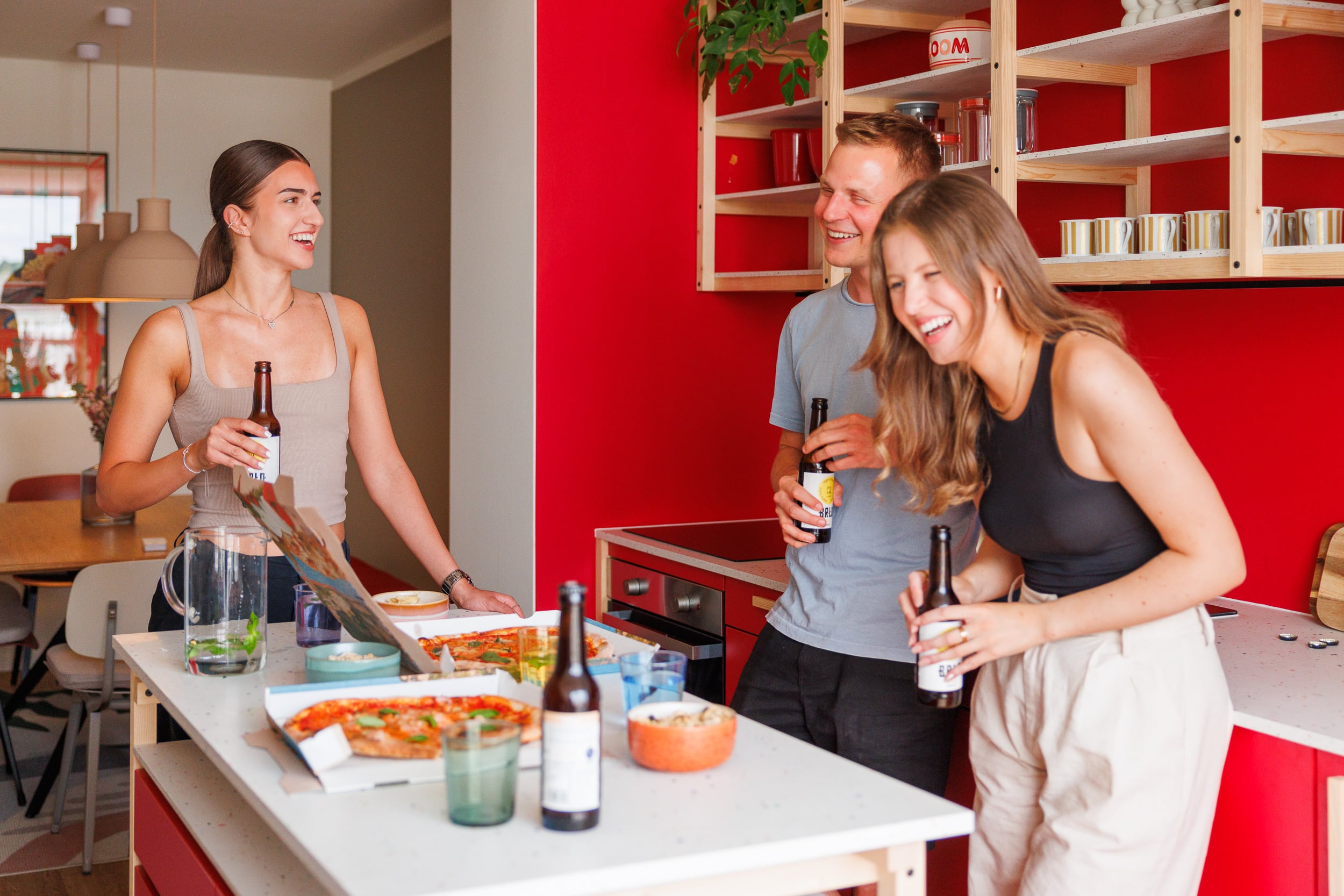 Three young adults laughing and holding drinks during a pizza party in a modern kitchen with a bright red wall.