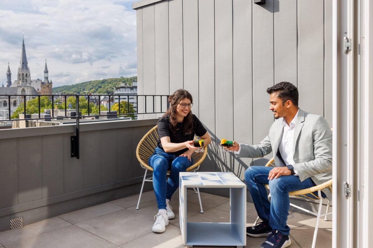 Man and woman enjoying drinks on a modern rooftop terrace overlooking the Aachen skyline and Cathedral on a sunny day.