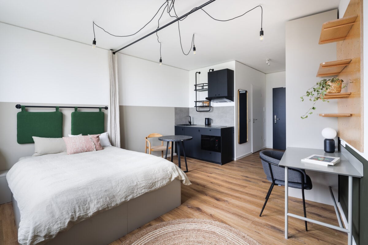 A modern minimalist studio apartment featuring a bed with green cushioned headboards, a small black kitchenette, a desk area, and hardwood floors.
