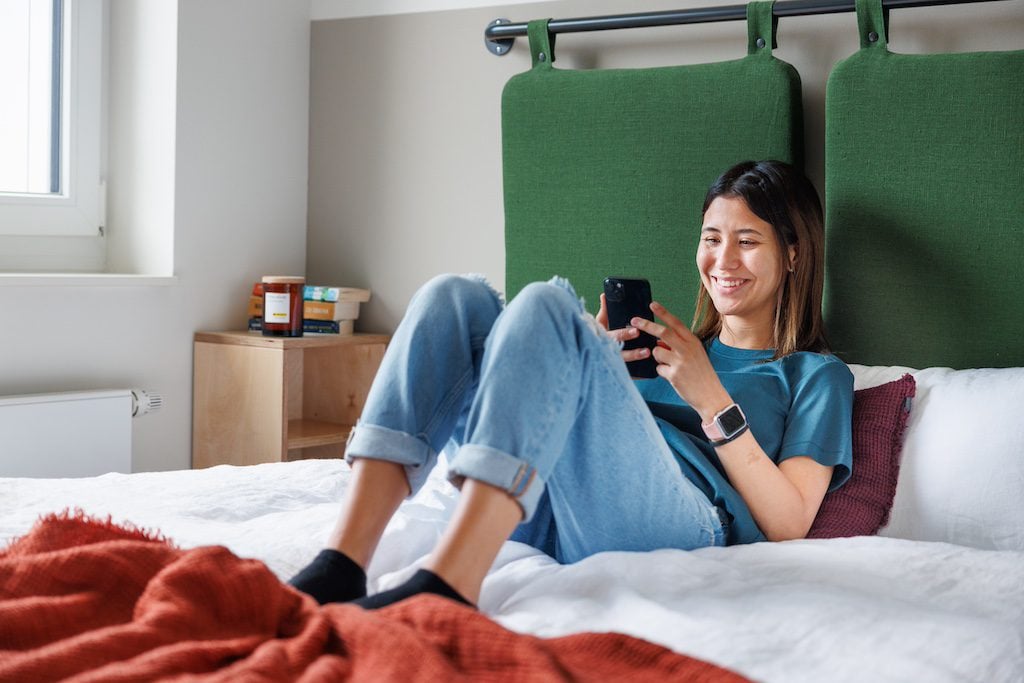 Frau sitzt auf einem Bett und lächelt, während sie auf ihr Smartphone schaut.