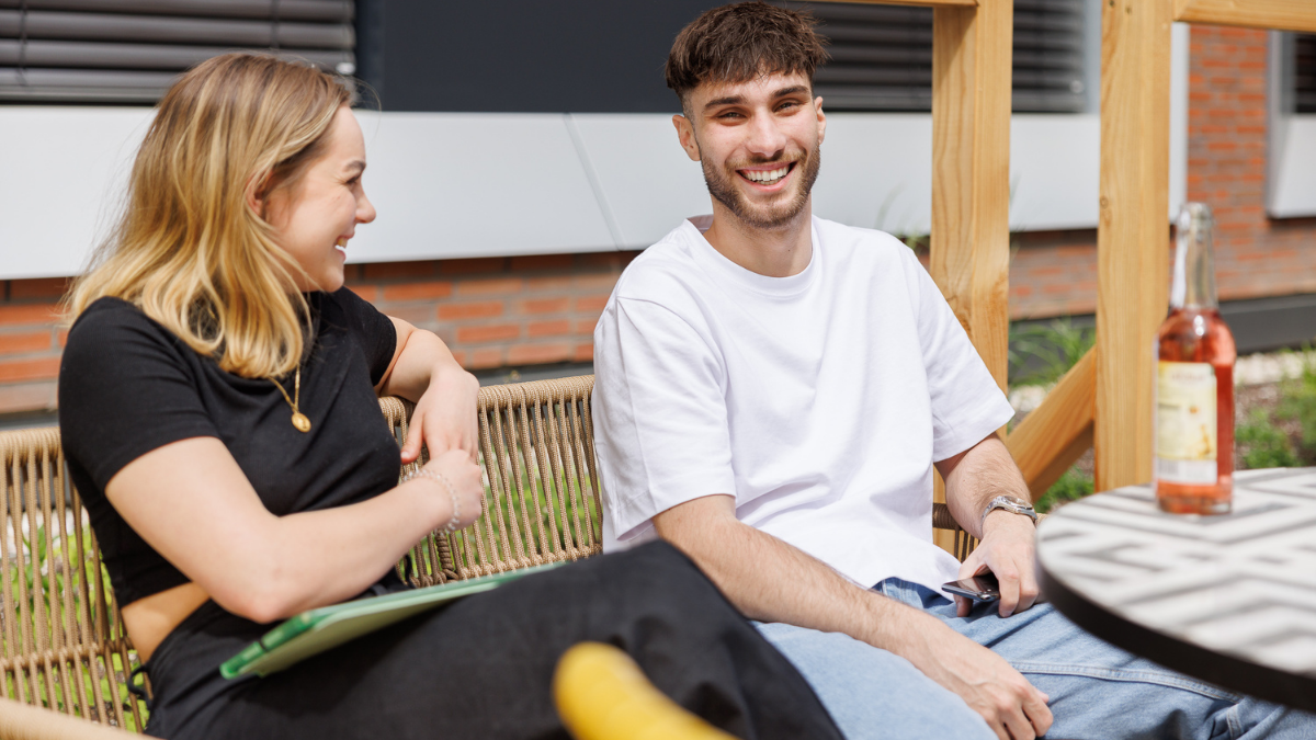 A young man in a white t-shirt smiling brightly while sitting outdoors with a young woman on a sunny day, suggesting friendship, community, or student life.