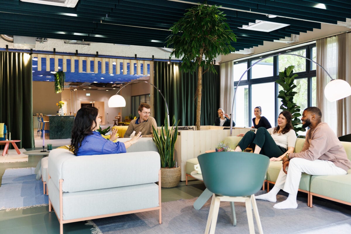 A group of diverse young professionals talking and laughing in a modern, brightly lit communal lounge with contemporary furniture, indoor plants, and large windows.