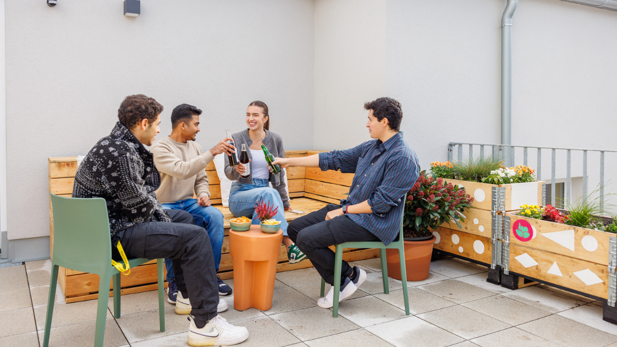 Four young adults are socializing on a bright outdoor terrace. They are sitting on wooden bench seating and modern green chairs, clinking glass bottles together in a toast. A small orange side table holds bowls of snacks. The setting is decorated with wooden planter boxes filled with flowers and greenery against a clean, light-colored building wall.