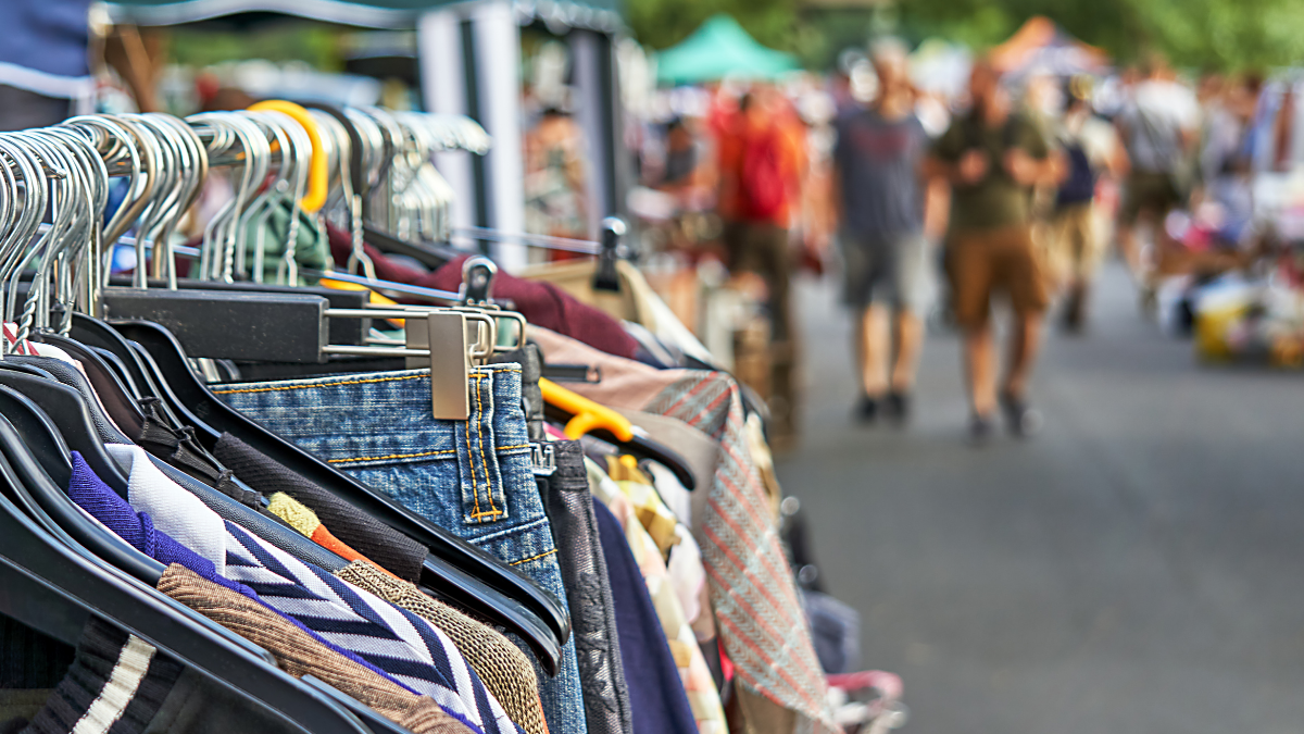 Close-up view of second-hand clothes (jeans, sweaters, jackets) hanging on racks at a bustling outdoor flea market, with blurred shoppers walking in the background.