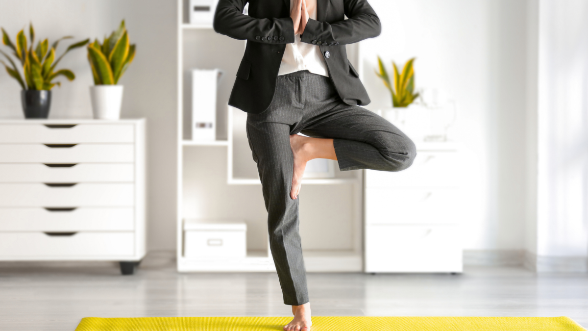 A person in business attire is barefoot and holding a tree pose (Vrksasana) on a yellow yoga mat in a home office. In the background, there are plants and white shelving units.