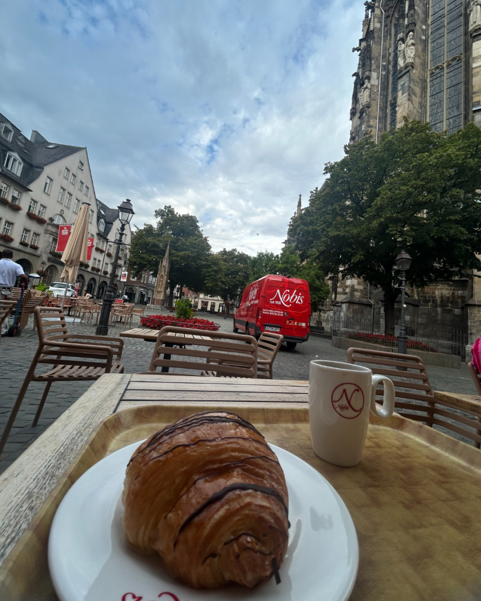 Chocolate croissant and coffee on an outdoor cafe table in Aachen, Germany, with the historic Cathedral visible in the background.