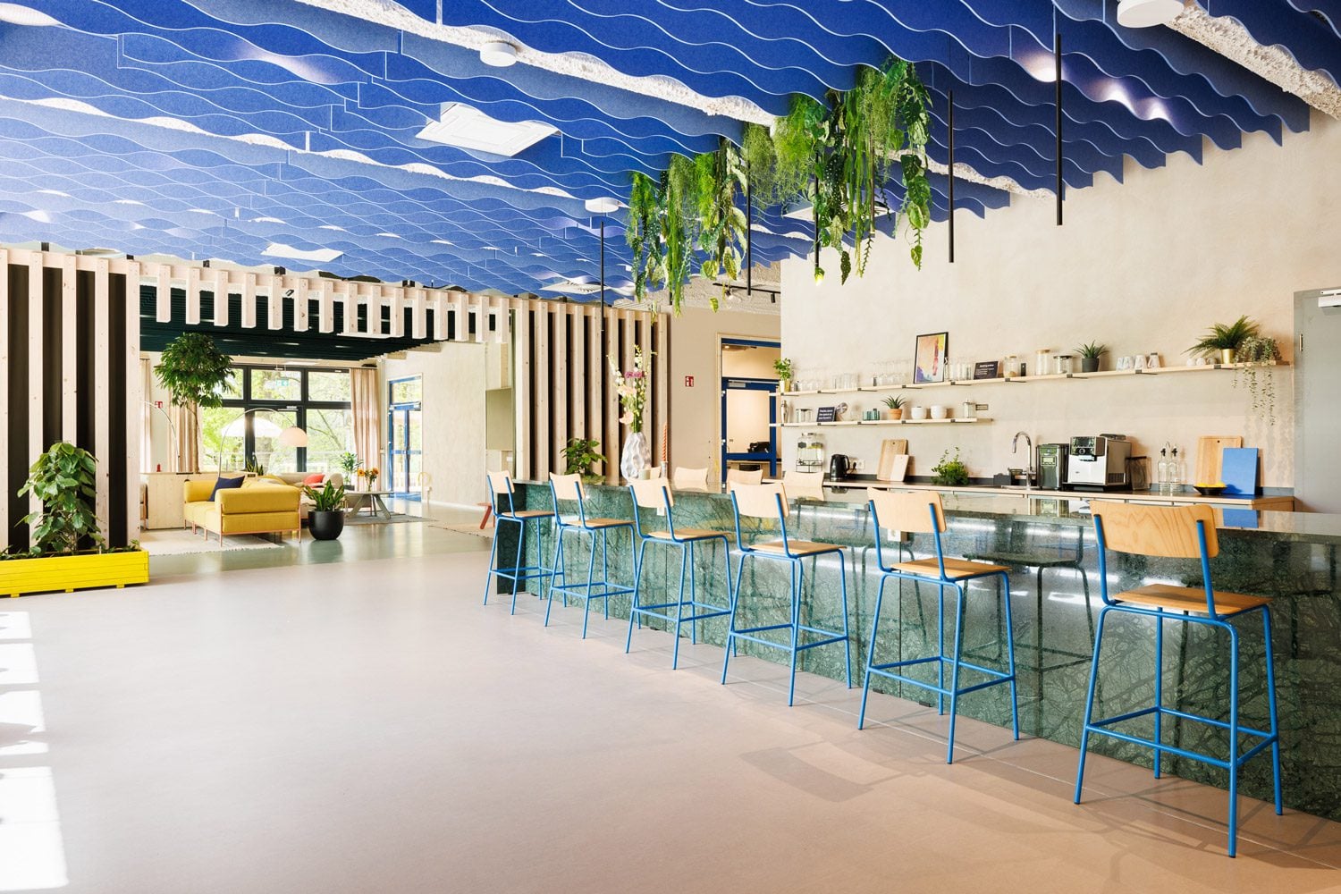 A modern open-plan office lounge and kitchen area featuring a wavy blue acoustic ceiling, a green marble breakfast bar with blue high chairs, and a bright seating area with a yellow sofa in the background.