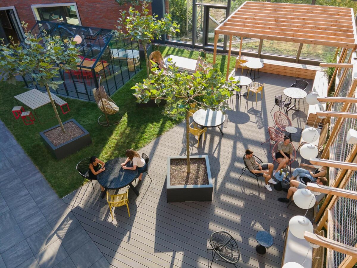 Eine Außenterrasse mit Tischen, Stühlen und Bäumen. Mehrere Personen sitzen und unterhalten sich in zwei Gruppen und genießen die Freiluft-Atmosphäre. Der Bereich verfügt über moderne Möbel und Grünflächen.