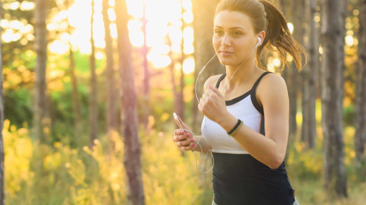 A young woman with a ponytail is jogging in a sunlit forest, wearing a black and white tank top and grey leggings. She has earbuds in and is holding a smartphone.