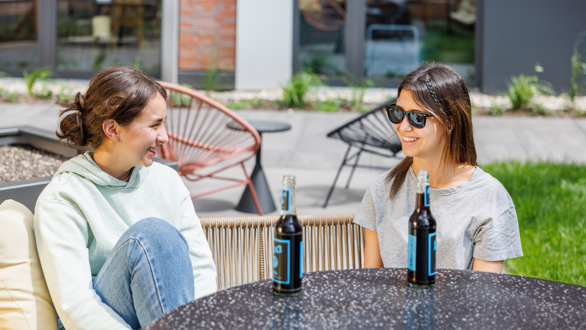 Two young women are sitting at a round, dark speckled table on an outdoor patio, laughing and talking together. One woman on the left wears a light-colored hoodie and jeans, while the woman on the right wears a grey t-shirt and dark sunglasses. Two glass soda bottles sit on the table between them. The background features modern outdoor furniture, including a red string chair, and a glimpse of a green lawn and building exterior.