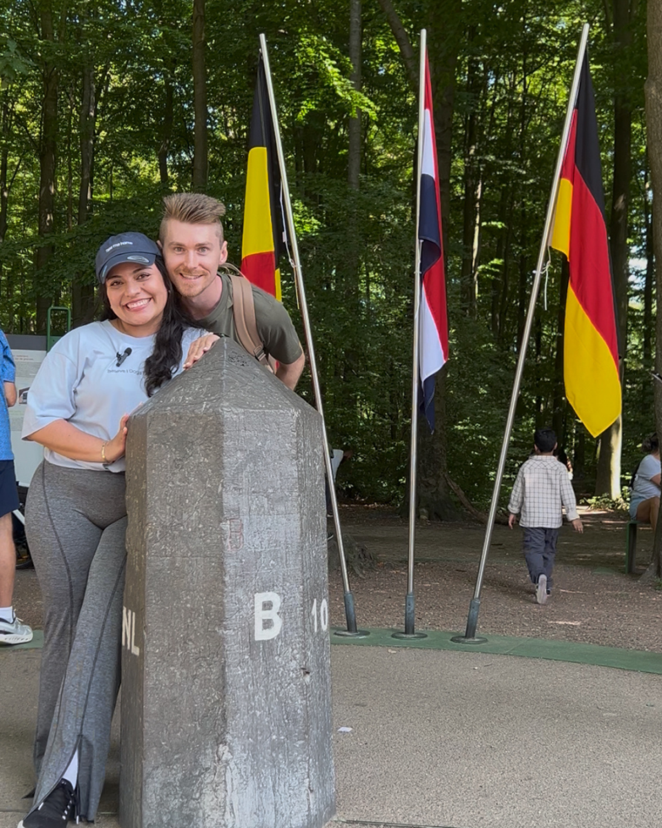 Close-up of a happy couple leaning against the gray stone tri-border marker engraved with NL and B, with the flags of three nations standing behind them in a forest.